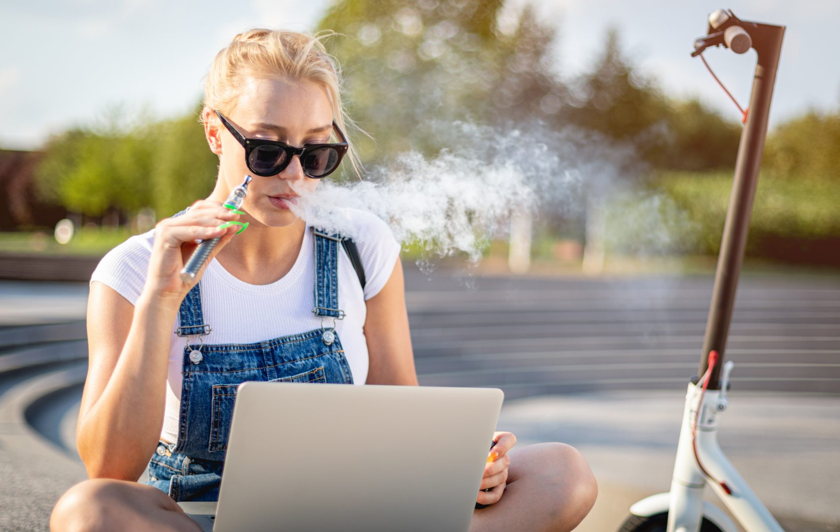 Modern woman smoking e-cigarette and using her laptop computer while sitting at street